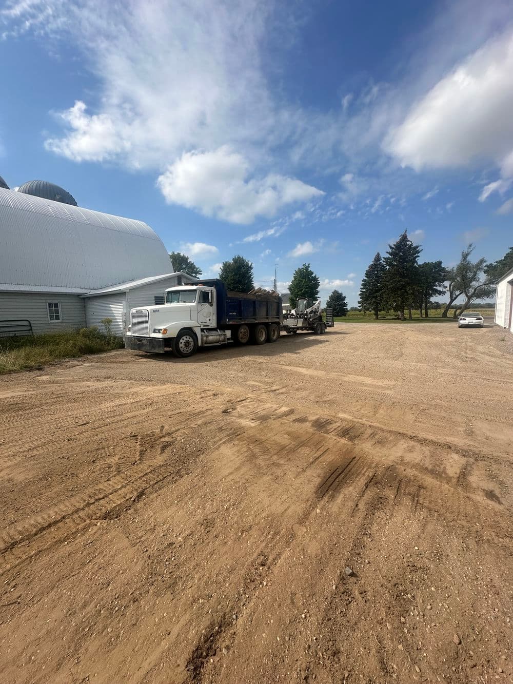 White dump truck parked on a gravel farmyard with trees and a barn in the background.