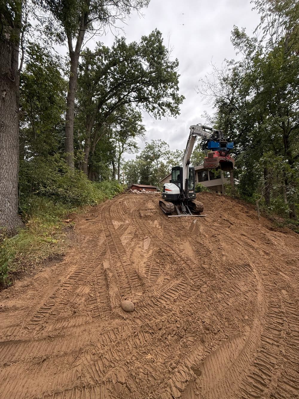 Excavator working on a dirt slope in a forested area with cloudy skies above.