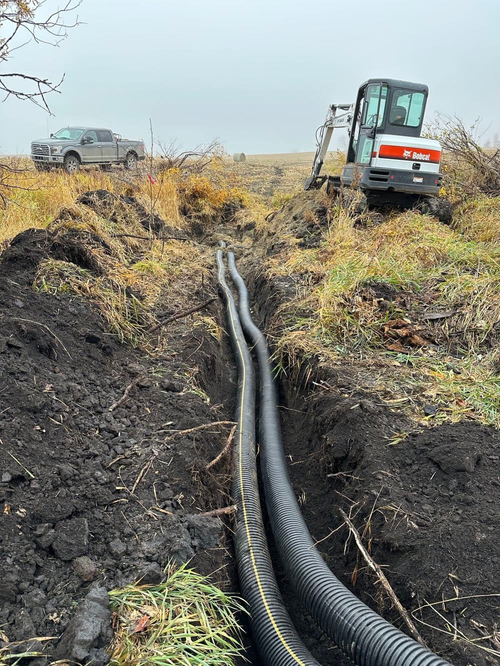Excavator installing black drainage pipes in a trench on a foggy field.