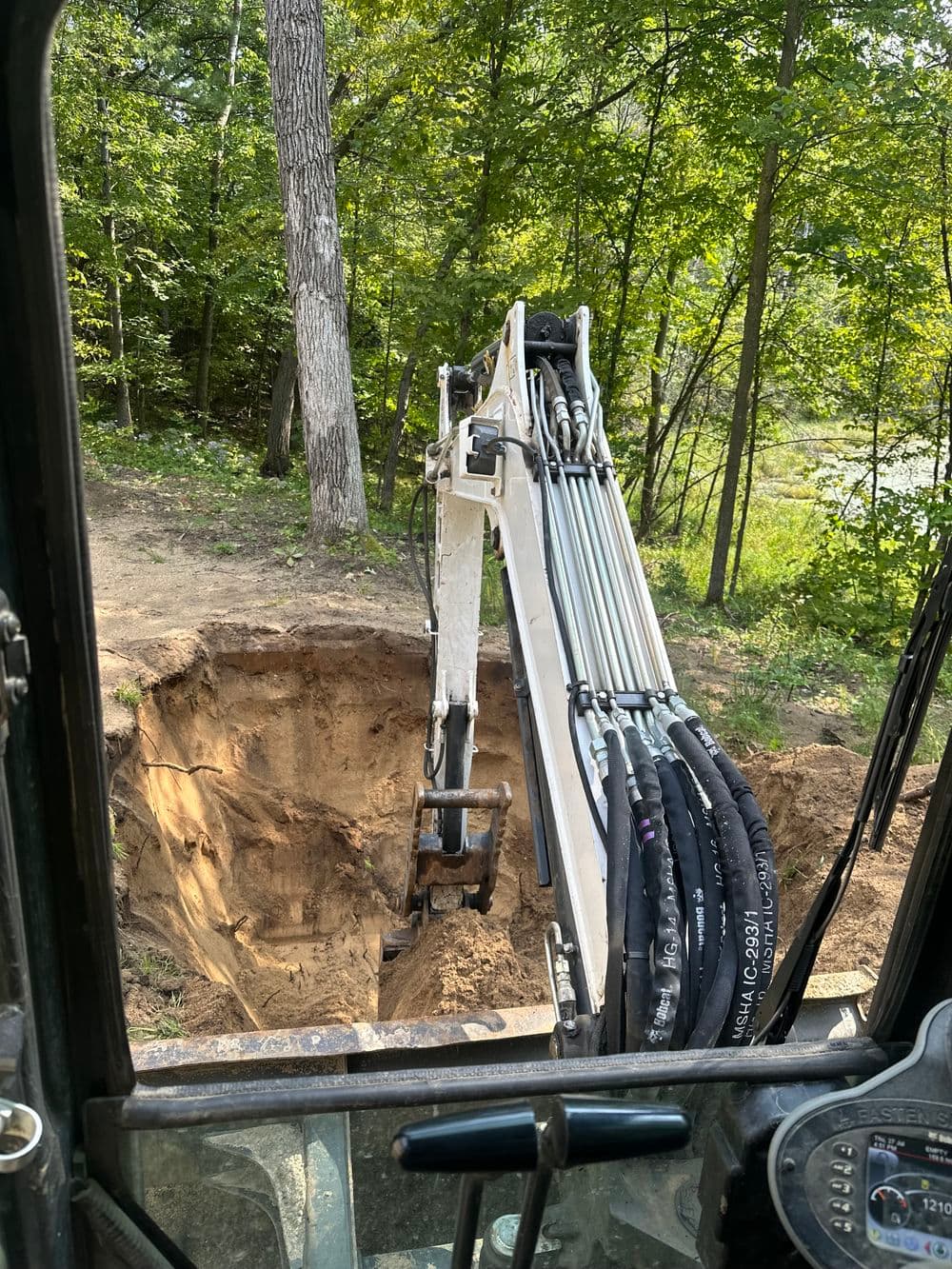 Excavator digging a large hole in a wooded area with trees in the background.
