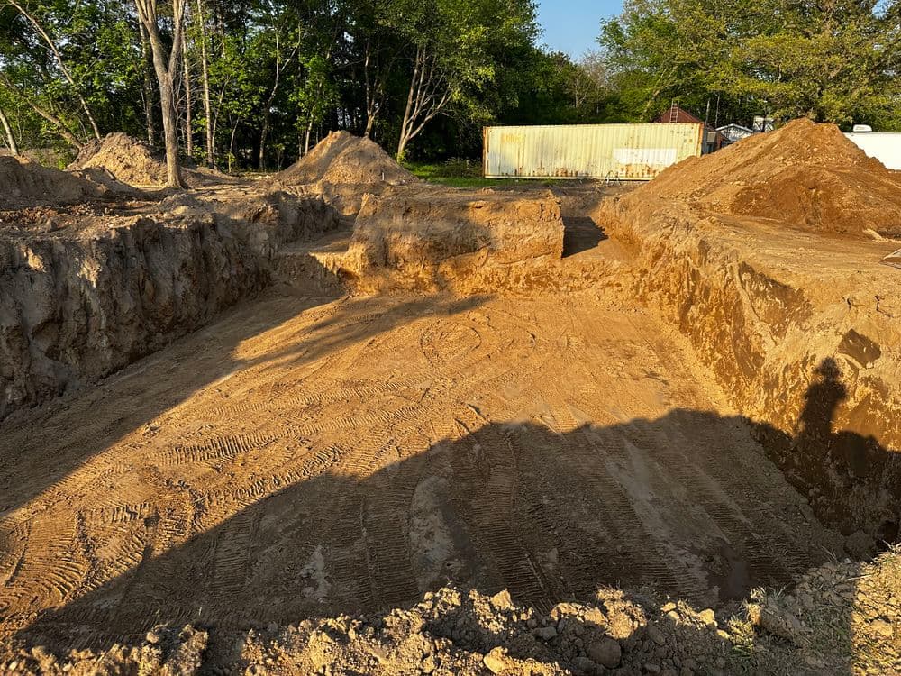 Excavated construction site with sandy soil, piles of dirt, and surrounding trees.