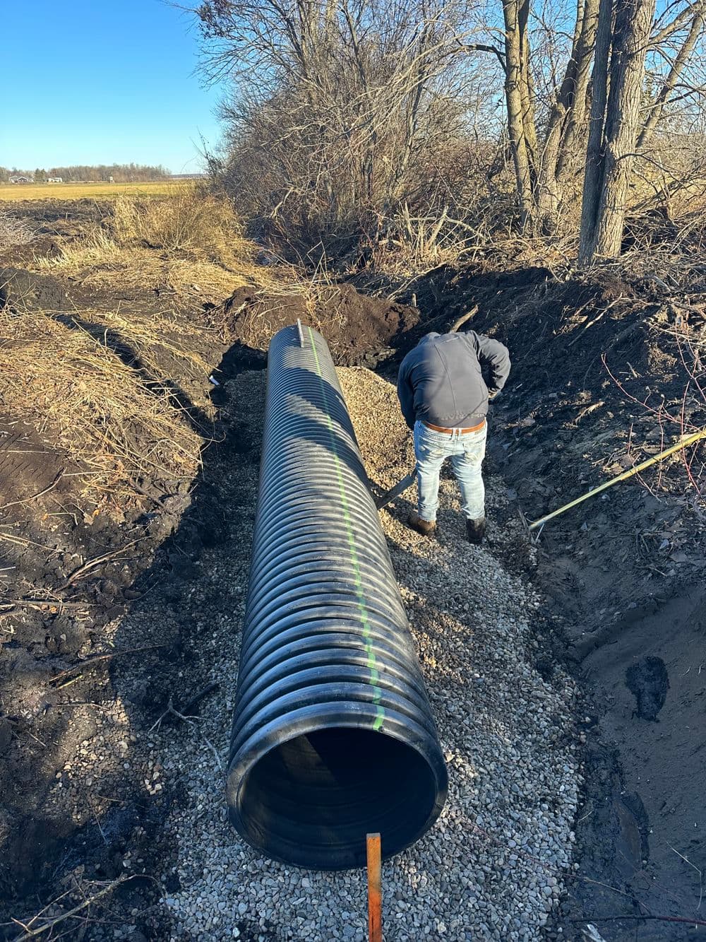 Worker installing a drainage pipe in a rural area surrounded by trees and grassland.
