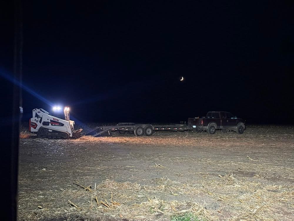 Nighttime farming scene with a skid steer loader and truck on a field under a crescent moon.
