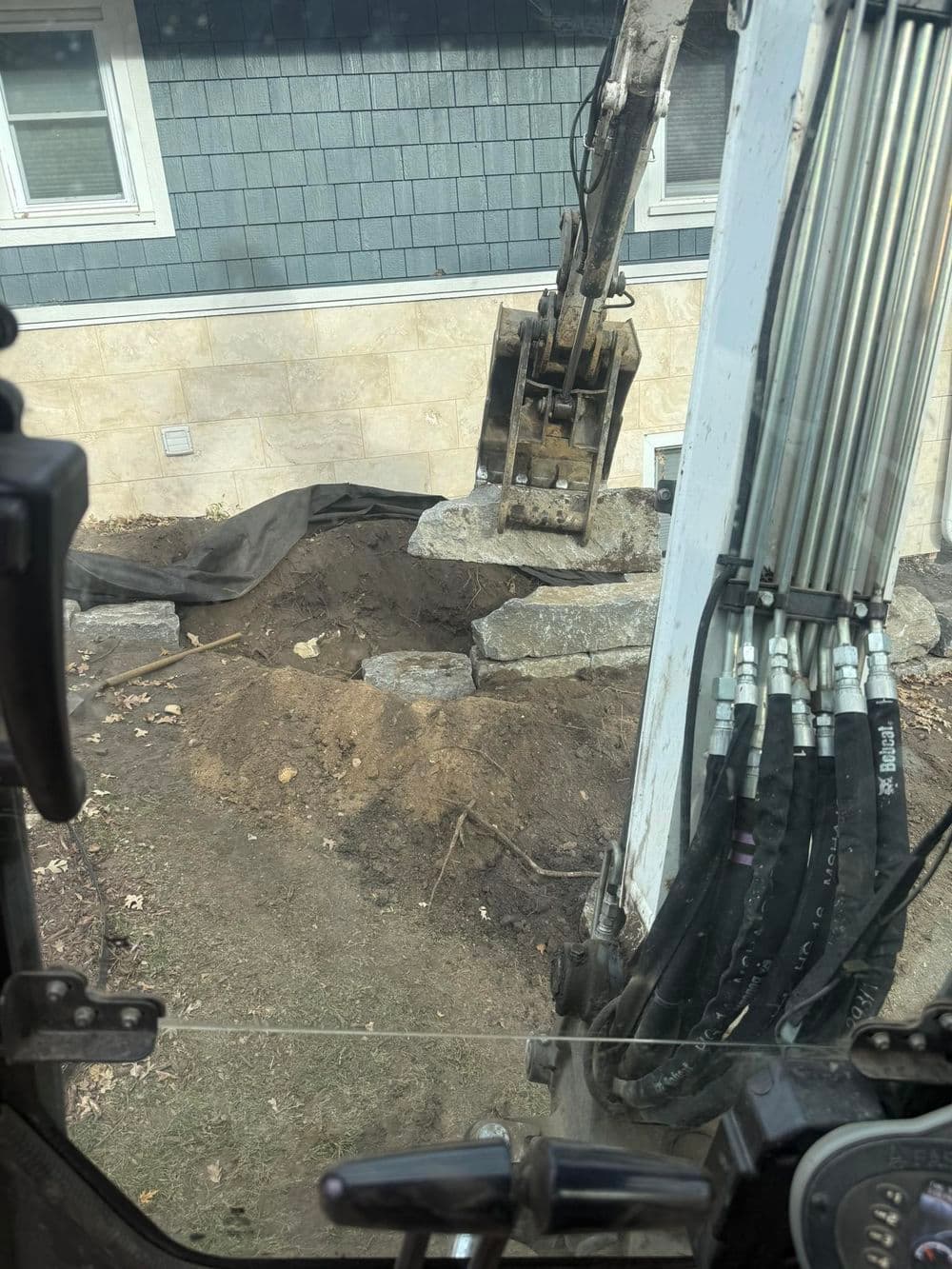Excavator bucket lifting stone near a home, with dirt and landscaping visible.