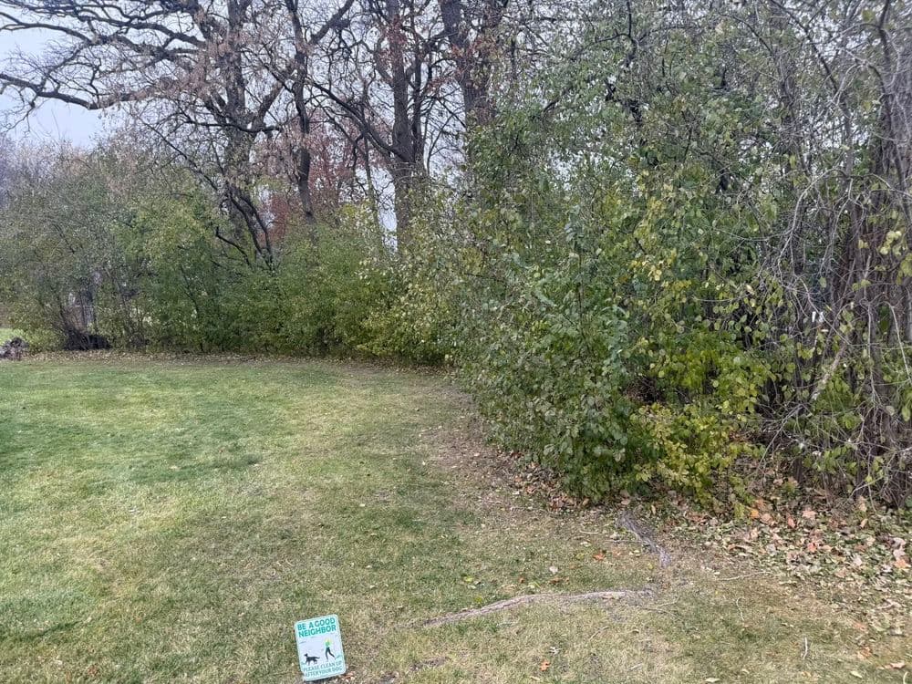 Dog waste disposal sign next to green grass and dense shrubs in a park setting.