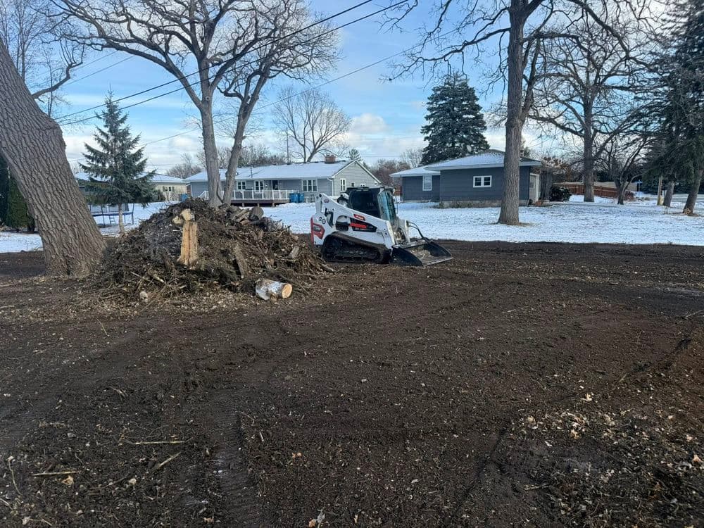 Bobcat skid steer clearing tree debris in snowy landscape with residential homes in background.