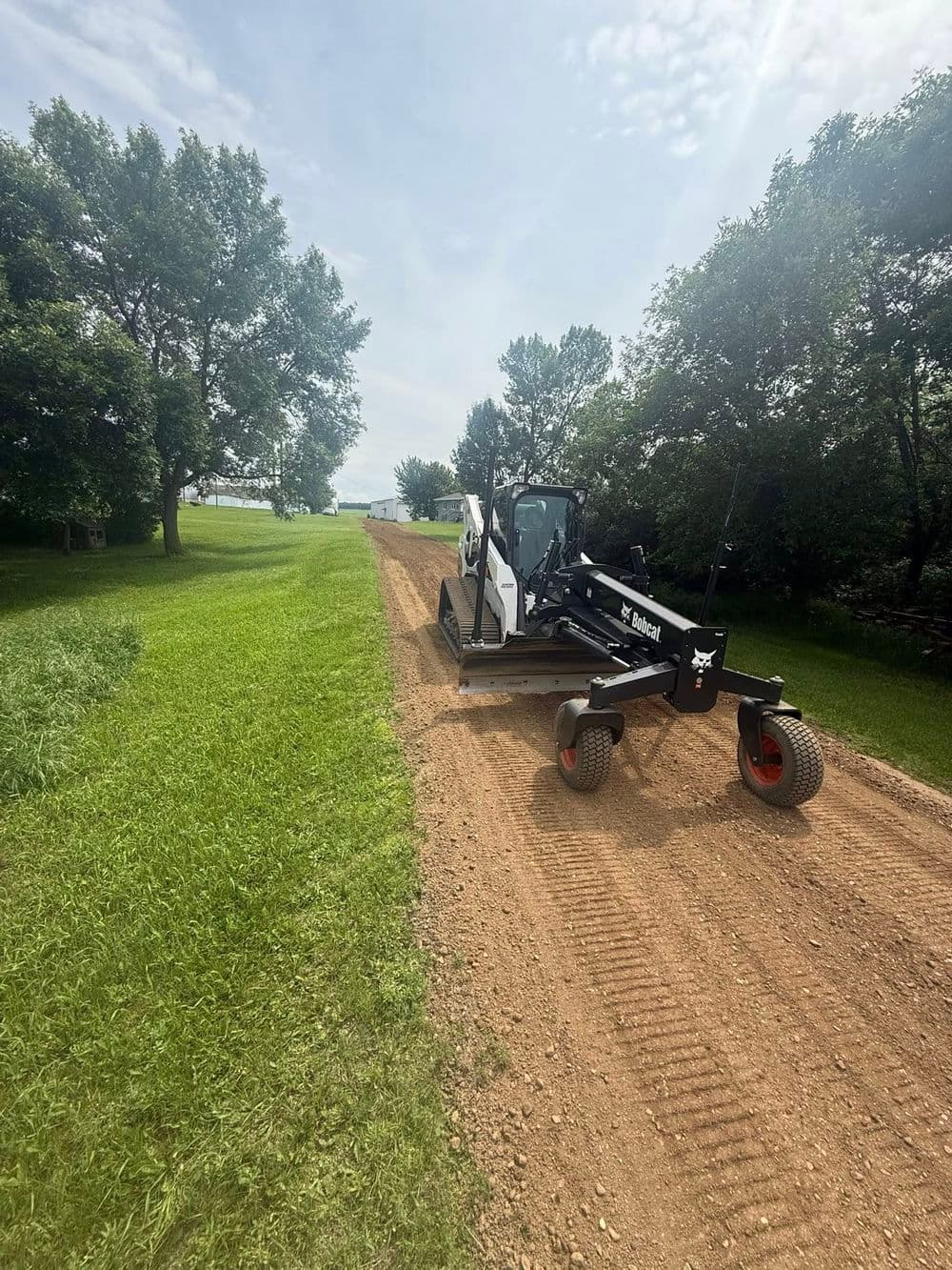 Bobcat grader leveling dirt road surrounded by greenery under a clear sky.