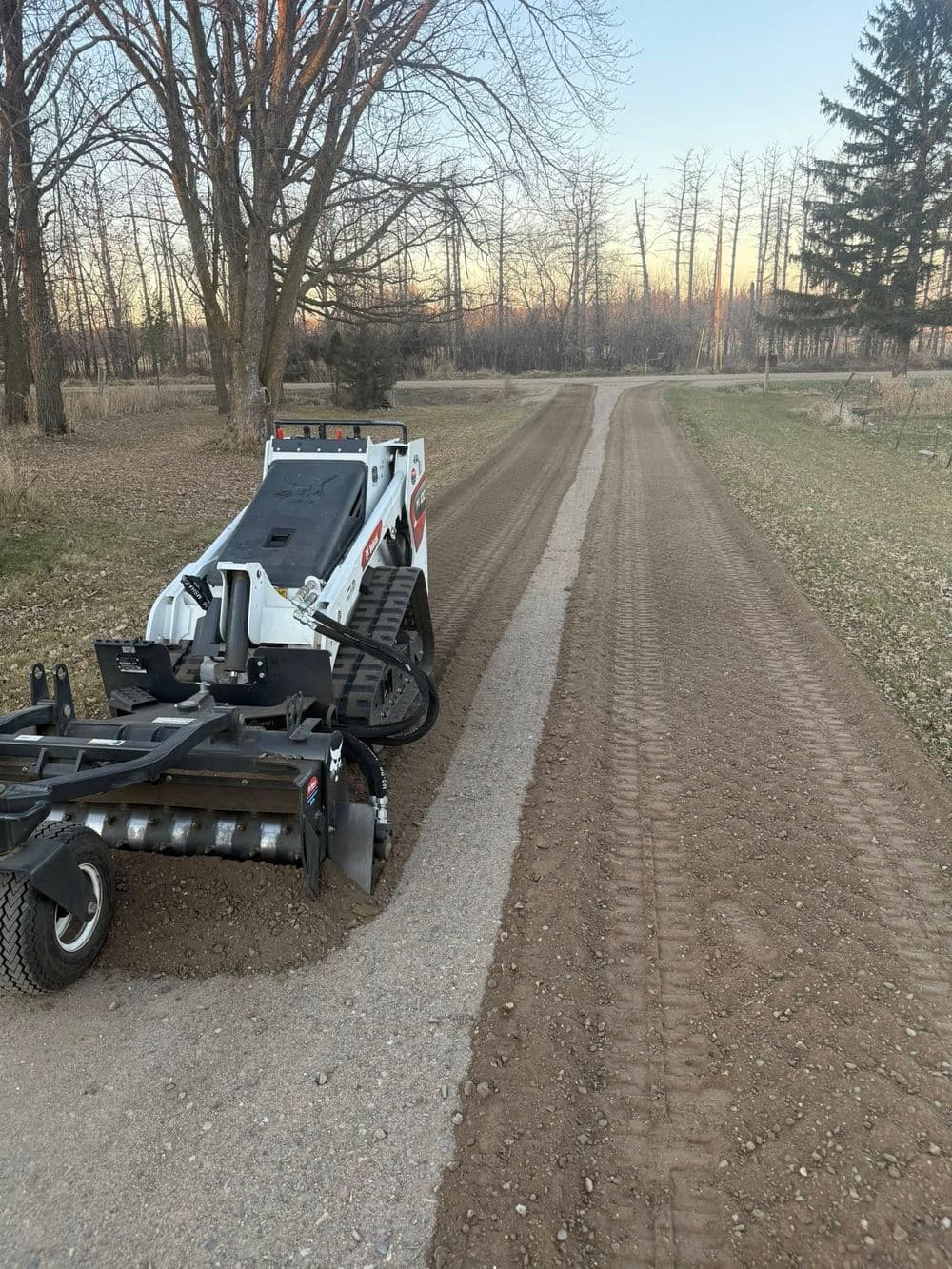 Bobcat grading machine on a rural dirt road, surrounded by trees at sunset.