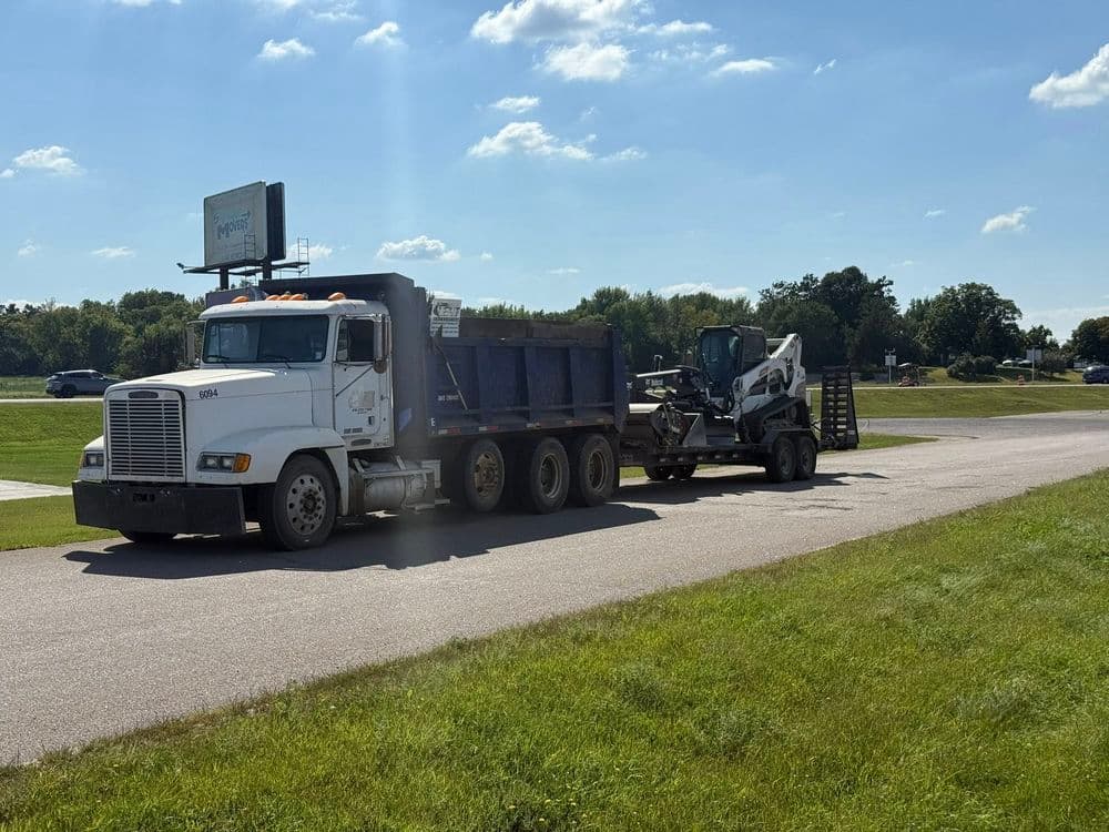 Dump truck and mini excavator parked on a roadside under a blue sky with clouds.